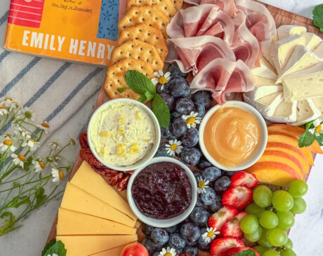 A colorful charcuterie board inspired by the cover of Beach Read by Emily Henry featuring brie, salami, peaches, blueberries, grapes, crackers, and edible flowers.