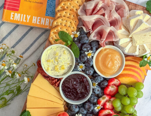 A colorful charcuterie board inspired by the cover of Beach Read by Emily Henry featuring brie, salami, peaches, blueberries, grapes, crackers, and edible flowers.