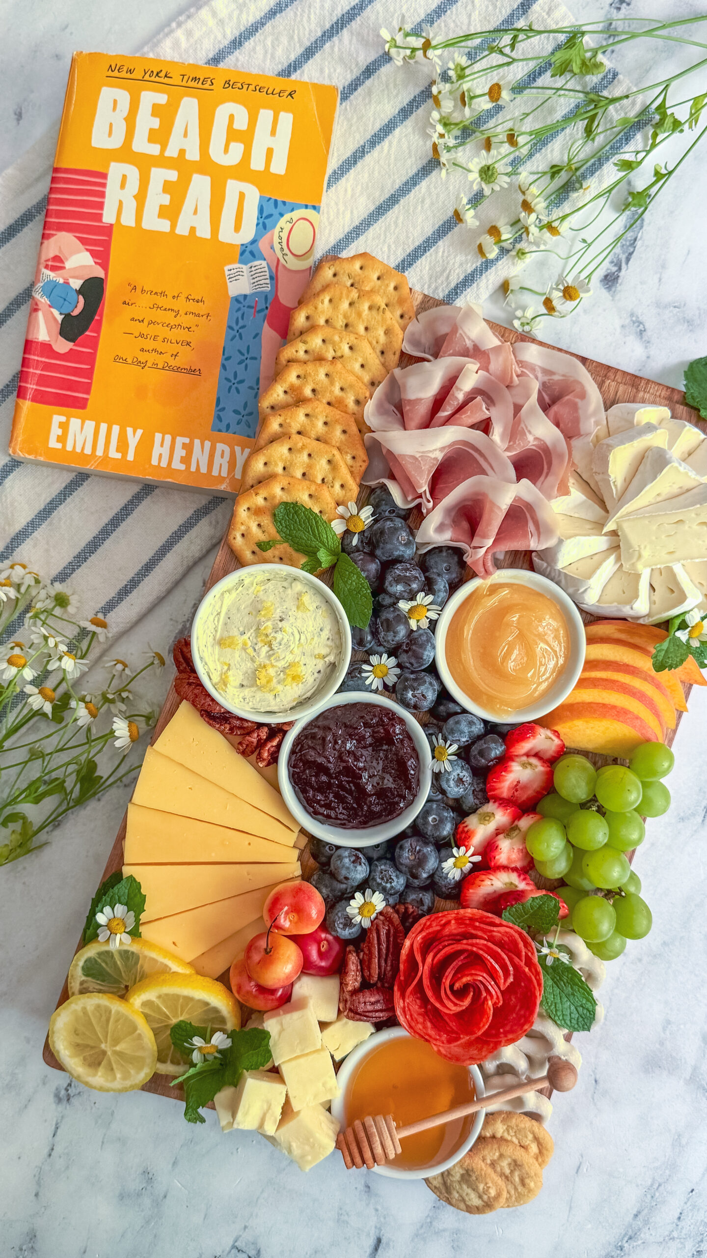 A colorful charcuterie board inspired by the cover of Beach Read by Emily Henry featuring brie, salami, peaches, blueberries, grapes, crackers, and edible flowers.